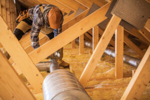 Professional HVAC Technician Installing Air Ducts in the Wooden Roof Section of the Building. Ventilation Systems Theme.
